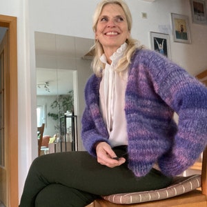 May include: A woman wearing a purple and pink striped cardigan with a white blouse and dark green trousers. She is sitting on a wooden chair with a striped cushion.