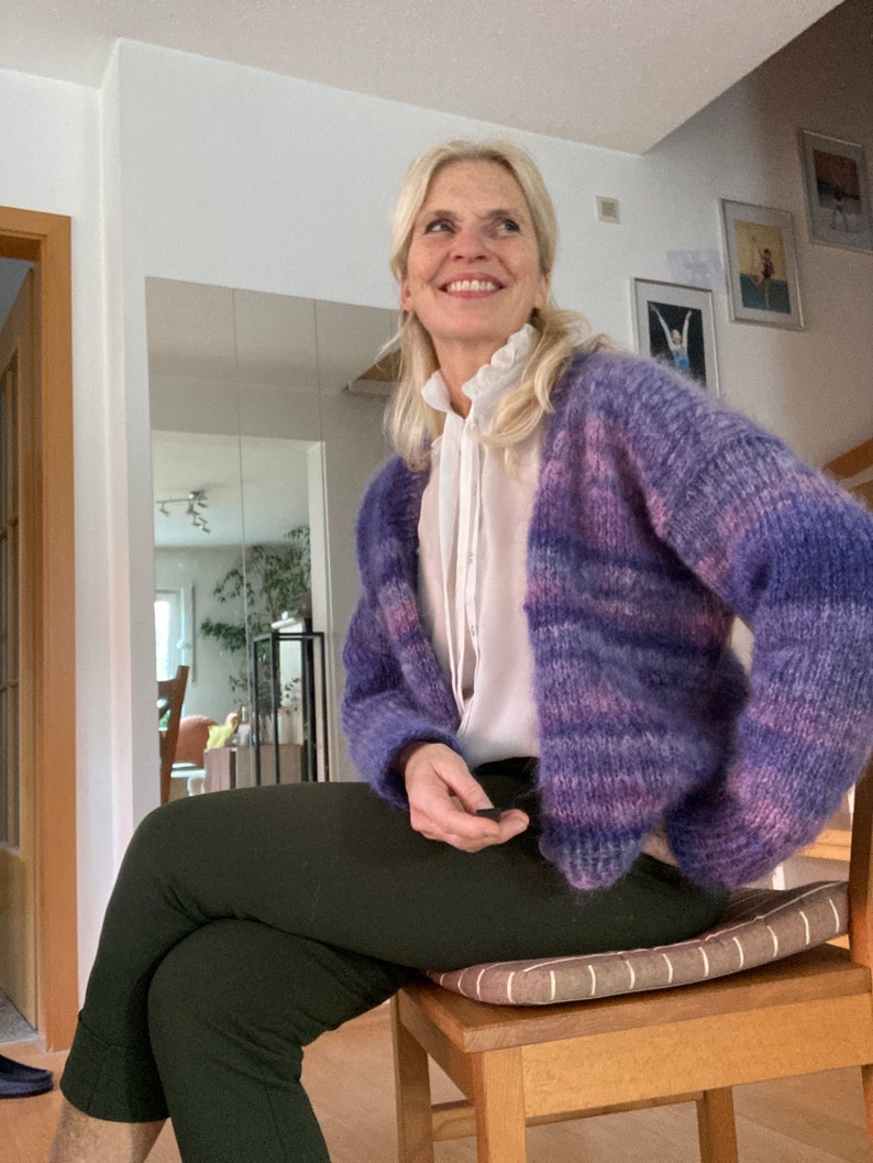 May include: A woman wearing a purple and pink striped cardigan with a white blouse and dark green trousers. She is sitting on a wooden chair with a striped cushion.