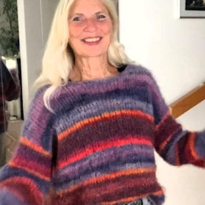 Könnte beinhalten: Ein mehrfarbiger gestreifter Pullover in Lila-, Rot- und Orangetönen. Der Pullover hat einen runden Ausschnitt und lange Ärmel. Die Frau trägt den Pullover mit einer grauen Jeans.