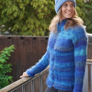 May include: A blue and teal striped fuzzy knit cardigan with a zip closure. The cardigan is being worn by a person wearing a blue knitted beanie.