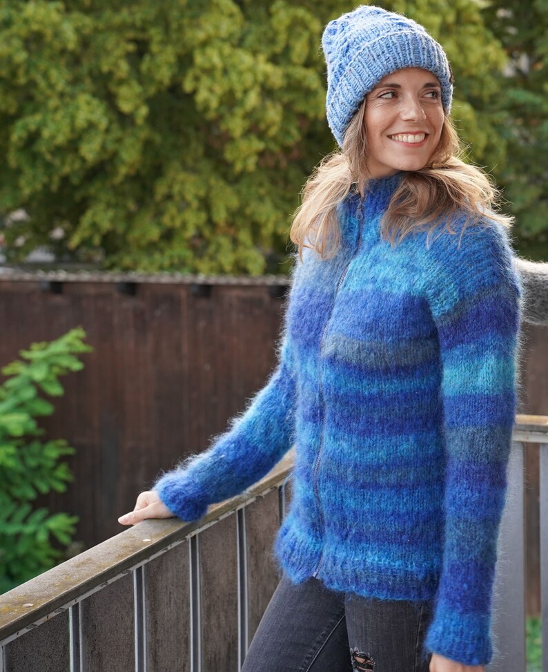 May include: A blue and teal striped fuzzy knit cardigan with a zip closure. The cardigan is being worn by a person wearing a blue knitted beanie.