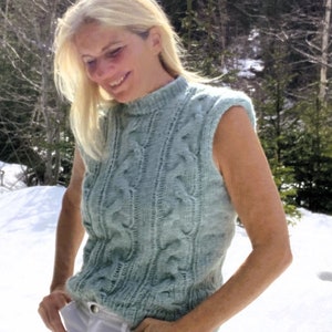 Puede incluir: Una mujer lleva un suéter sin mangas de punto azul claro con cuello alto blanco. Está de pie en un paisaje nevado.