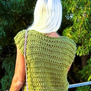 Può includere: Un top in maglia verde oliva a maniche corte, caratterizzato da una struttura a maglia larga e aperta con un ampio scollo. Il capo presenta un aspetto lavorato a mano con colonne verticali visibili di asole di filato.