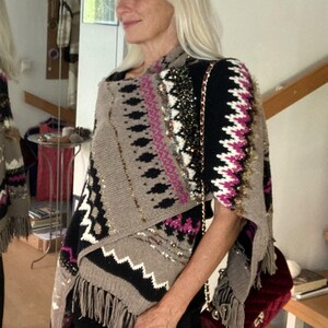 May include: A knit poncho featuring a geometric pattern in beige, black, white, and magenta. The garment includes metallic gold thread accents, sequin embellishments, and a fringed hemline. It is worn over black clothing with a dark red velvet bag.
