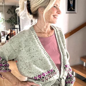 Op de afbeelding: Lichtgroen vest met een bloemenpatroon in paars en zwart op de mouwen en zoom. Het vest wordt gedragen over een roze top en een olijfgroene broek. De vrouw draagt een bruine haarclip.