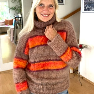 Könnte beinhalten: Ein brauner, übergroßer Pullover mit Rollkragen und horizontalen Streifen in Orange- und Rottönen. Der Pullover hat lange Ärmel und ein strukturiertes, flauschiges Aussehen, was auf ein warmes Strickdesign hindeutet. Der Gesamtstil ist lässig und bequem.