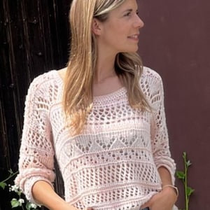 Könnte beinhalten: Ein hellrosa, offen gestrickter Pullover mit Rundhalsausschnitt und Dreiviertelärmeln. Der Pullover hat ein strukturiertes, gemustertes Design. Die Person trägt eine blaue Jeans. Der Hintergrund ist ein dunkler Holzzaun und eine weinrote Wand.