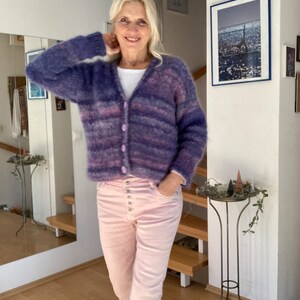 May include: A woman wearing a purple and pink striped cardigan with a white shirt underneath and pink trousers. She is standing in a room with a mirror and a picture on the wall.