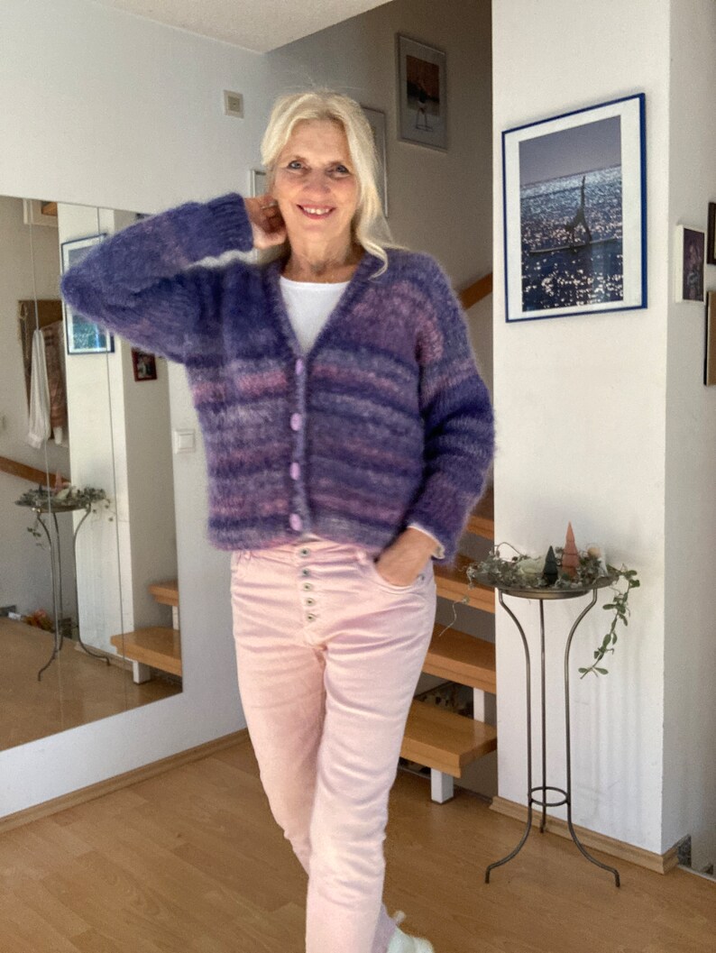 May include: A woman wearing a purple and pink striped cardigan with a white shirt underneath and pink trousers. She is standing in a room with a mirror and a picture on the wall.