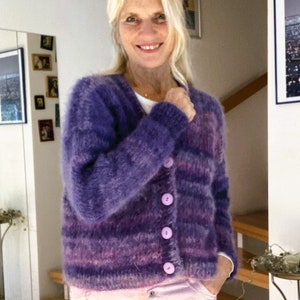 May include: A purple and pink striped cardigan with a fuzzy texture and five light purple buttons. The cardigan is worn over a light pink shirt.