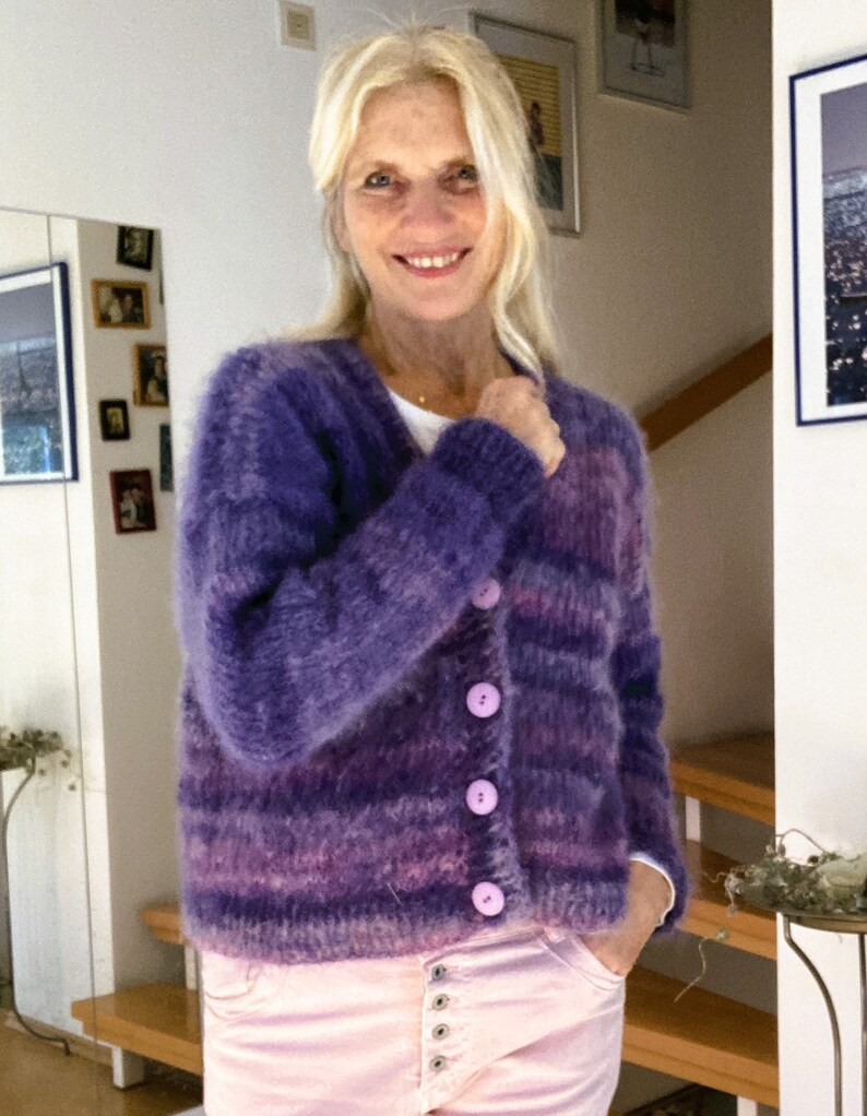 May include: A purple and pink striped cardigan with a fuzzy texture and five light purple buttons. The cardigan is worn over a light pink shirt.