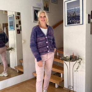 May include: A woman wearing a purple and pink striped cardigan with a white shirt underneath and pink trousers. She is standing in a room with a wooden floor and a staircase in the background.