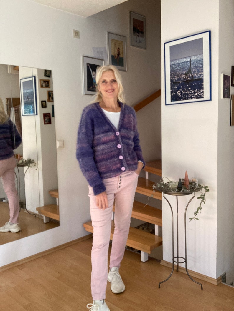 May include: A woman wearing a purple and pink striped cardigan with a white shirt underneath and pink trousers. She is standing in a room with a wooden floor and a staircase in the background.
