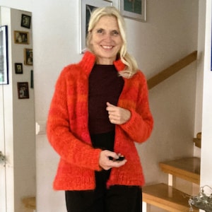 May include: A woman wearing a bright orange and red fuzzy knit cardigan with a dark brown turtleneck shirt underneath. She is standing in front of a white wall with a wooden staircase behind her.