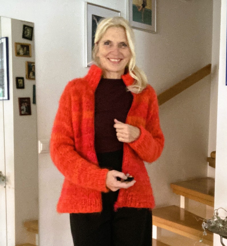 May include: A woman wearing a bright orange and red fuzzy knit cardigan with a dark brown turtleneck shirt underneath. She is standing in front of a white wall with a wooden staircase behind her.