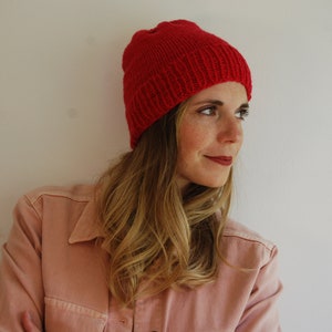 May include: A woman wearing a red knit beanie hat and a pink collared shirt.