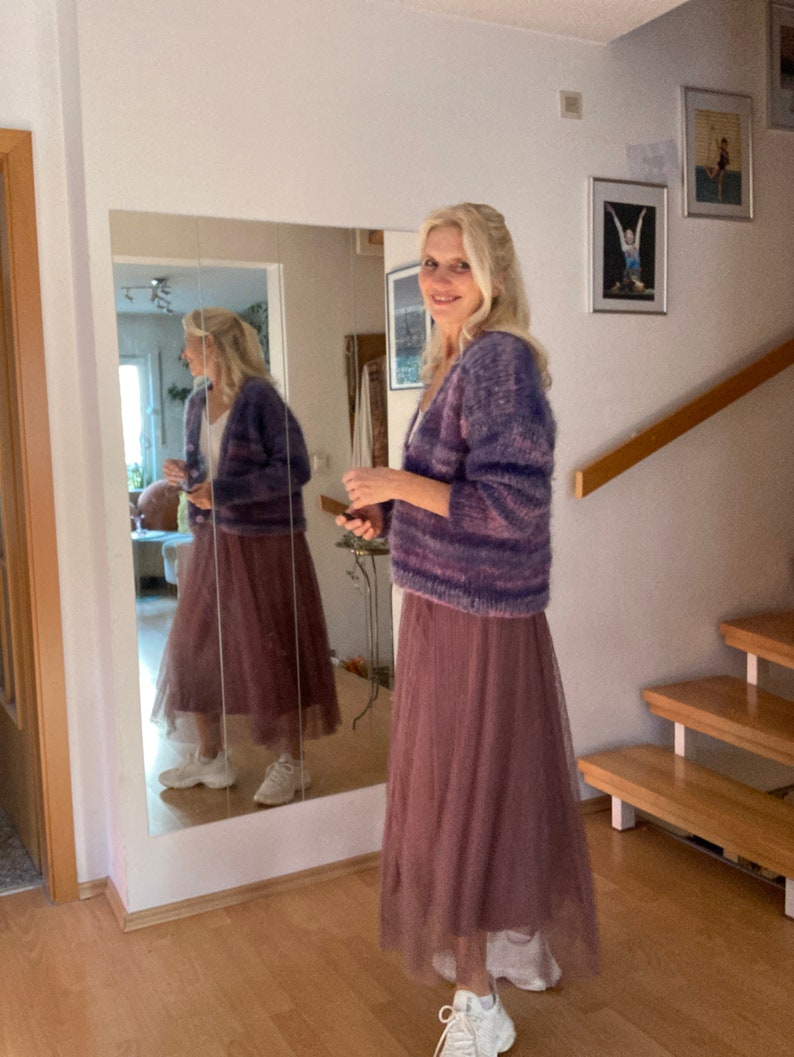 May include: A woman wearing a purple and pink striped cardigan with a long, dusty rose tulle skirt and white trainers.