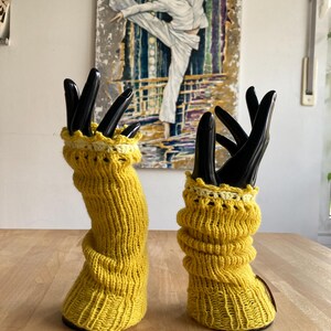 May include: A pair of yellow knitted arm warmers with white crocheted trim. The arm warmers are displayed on a pair of black mannequin hands.