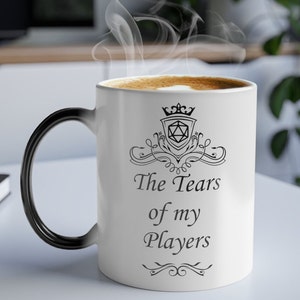 Peut inclure: Une tasse à café en céramique blanche avec une poignée noire. La tasse présente un motif noir d'une couronne, d'un dé à 20 faces et le texte "The Tears of my Players".