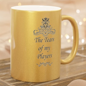 May include: Gold mug with a black and white design featuring a crown, a dice, and the text "The Tears of my Players".