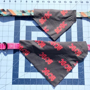 May include: Two dog collars with bandanas. One collar has a geometric pattern in teal, orange, and black with a gold buckle. The other is pink. Both bandanas are black with the red text "AC/DC".