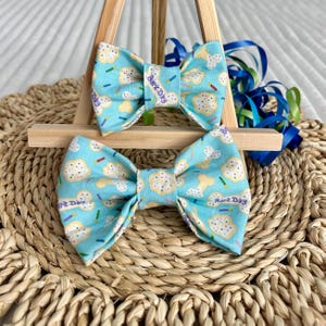 May include: Two blue bow ties with a cookie pattern and the text "Bark Day" on them. The bow ties are on a wooden stand and a woven mat.