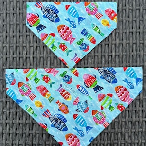 May include: Two blue dog bandanas with a colorful fish pattern. The fish are in various colors, including red, orange, green, and blue, with intricate designs. The bandanas are triangular in shape.