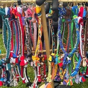 Op de afbeelding: Een display van kleurrijke, gevlochten hondenriemen. De riemen zijn gemaakt van verschillende stoffen in tinten rood, blauw, groen en geel. Elke riem is versierd met stoffen accenten en lussen voor bevestiging aan de halsband.