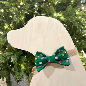May include: A green bow tie with a white background and a pattern of colorful Christmas lights. The bow tie is tied around a wooden dog head cutout.