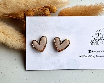 May include: A pair of gold heart-shaped earrings with a speckled white and beige design. The earrings are displayed on a white card with the text "Karo & Clay Jewellery" printed on it.