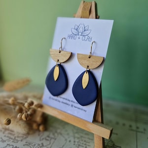 May include: Earrings on a wooden display stand. Each earring has a wooden semi-circle top, a navy blue teardrop shape, and a gold-coloured leaf detail. The earrings are presented on a card with the text "KARO & CLAY" and a lotus flower logo.