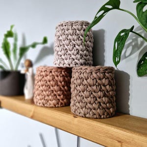 May include: Three crocheted plant pots in different shades of brown and beige. The pots are stacked on a wooden shelf.