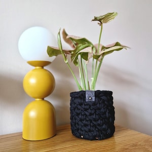 May include: A black crocheted plant pot with a "HAND MADE" label, holding a pink and green houseplant. A yellow, three-tiered lamp sits on a wooden surface next to the plant.
