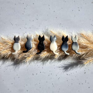 May include: Six cat-shaped ornaments in a row, each with a unique colour scheme. The cats are white, grey, and black, with speckled and gradient designs. They are arranged on a bed of dried, light brown grass against a light grey background.