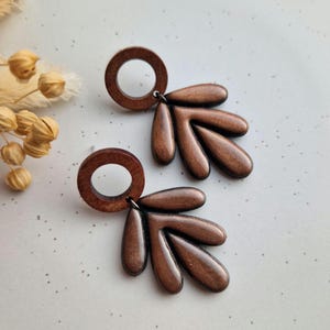 May include: A pair of brown earrings with a circular top and a leaf-like design. The earrings are made of a dark brown material with a metallic sheen. The earrings are hanging on a white surface.