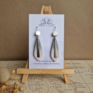 May include: Handmade clay earrings with a teardrop design, displayed on a miniature easel. The earrings feature a gradient of colours, transitioning from pale pink to grey. They have silver-coloured metal hooks. The card behind the earrings has the text "KARO & CLAY".
