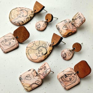 May include: A set of six pairs of earrings featuring abstract line drawings of faces on a peach background. The earrings are made of resin and wood and come in a variety of shapes, including teardrops, squares, and circles.