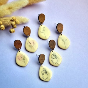 Peut inclure: Boucles d'oreilles artisanales avec une partie supérieure en bois et des pendentifs en forme de goutte d'eau crème. Chaque pendentif est orné d'un dessin au trait noir délicat d'un visage. Présentées sur une surface violet clair.
