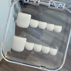 Könnte beinhalten: Eine transparente Kunststoffbox enthält zwei Reihen weißer künstlicher Zehennägel. Jede Reihe hat einen größeren Nagel und vier kleinere, alle mit glänzendem Finish. Die Nägel sind so gestaltet, dass sie einem natürlichen Fuß ähneln.
