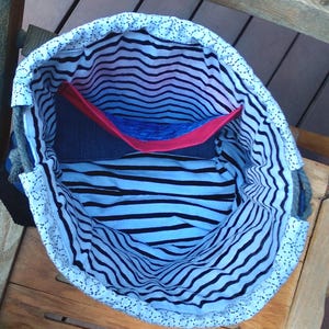 May include: Open denim tote bag with a black and white striped interior. The bag has a red and blue pocket. The top edge is white with a dotted pattern. The bag is sitting on a wooden surface.