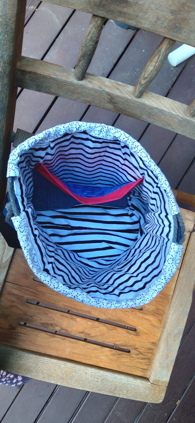 May include: Open denim tote bag with a black and white striped interior. The bag has a red and blue pocket. The top edge is white with a dotted pattern. The bag is sitting on a wooden surface.