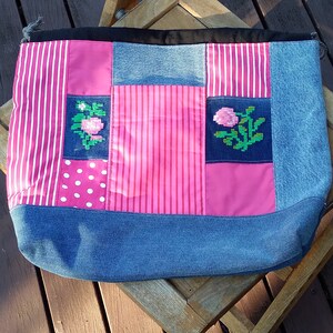 Op de afbeelding: Een patchwork tote bag met een denim basis en diverse roze en wit gekleurde stoffen. De tas heeft geborduurde roos details en een zwarte bovenrand. De tas ligt op een houten stoel.