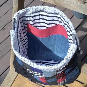 May include: Handmade patchwork tote bag with a denim exterior, red pocket, and striped lining. The bag sits on a wooden chair. The bag's design incorporates various fabric patterns and colors, creating a unique, handcrafted aesthetic.