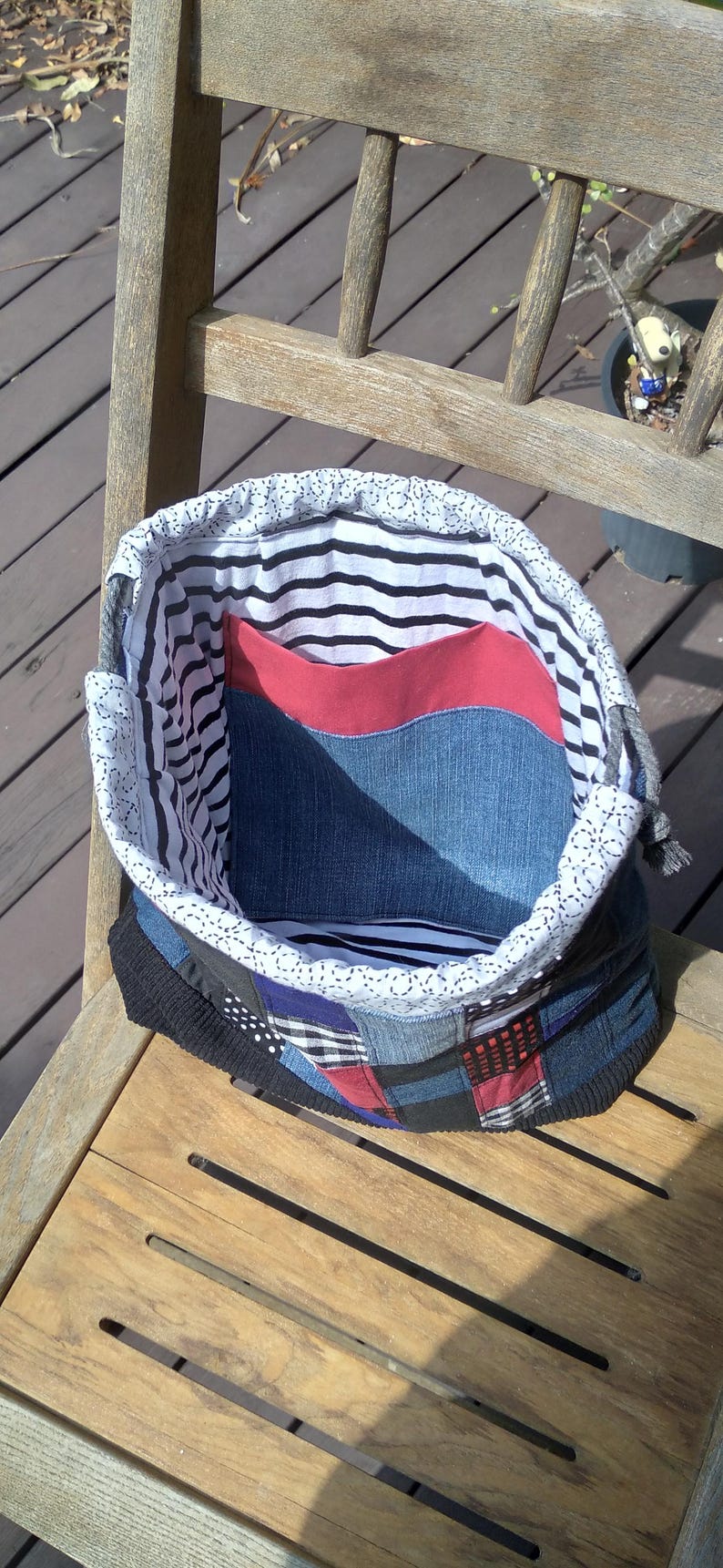 May include: Handmade patchwork tote bag with a denim exterior, red pocket, and striped lining. The bag sits on a wooden chair. The bag's design incorporates various fabric patterns and colors, creating a unique, handcrafted aesthetic.