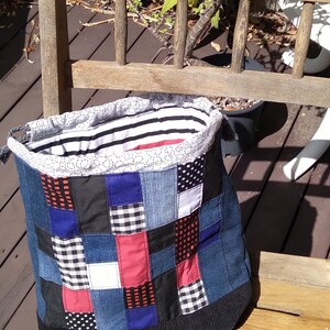 May include: A patchwork denim tote bag with a black knit bottom and a striped lining. The bag features squares of denim, red, black, white, and blue fabric. The bag is sitting on a wooden bench.