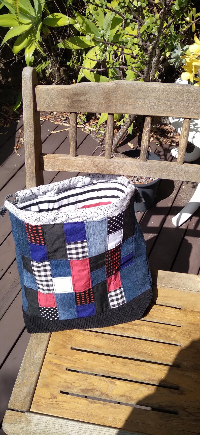 May include: A patchwork denim tote bag with a black knit bottom and a striped lining. The bag features squares of denim, red, black, white, and blue fabric. The bag is sitting on a wooden bench.