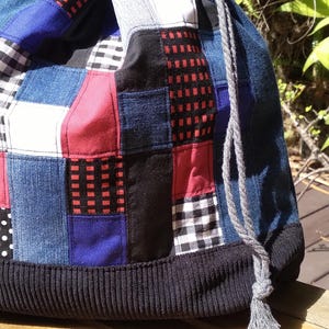 May include: A patchwork drawstring bag with a variety of fabric patterns and colors. The bag features denim, red, black, white, and blue squares. A black corduroy band forms the base, and a gray rope with a tassel serves as the drawstring.