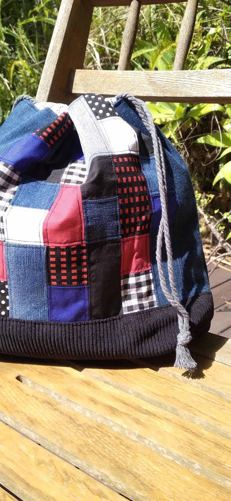 May include: A patchwork drawstring bag with a variety of fabric patterns and colors. The bag features denim, red, black, white, and blue squares. A black corduroy band forms the base, and a gray rope with a tassel serves as the drawstring.