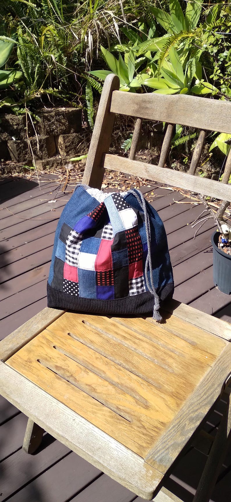 May include: A patchwork denim drawstring bag with a black knit base. The bag features squares of denim, red, white, black, and blue fabrics. It sits on a wooden chair on a deck.