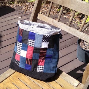 May include: A patchwork tote bag with a denim body and a black knit bottom sits on a wooden chair. The patchwork design includes squares of blue denim, red, black, and white patterned fabrics. The bag's top is a light-colored fabric with a small white pattern.